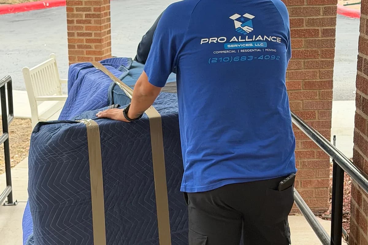 Pro Alliance mover moving a blanket-wrapped sofa on a dolly along an exterior walkway