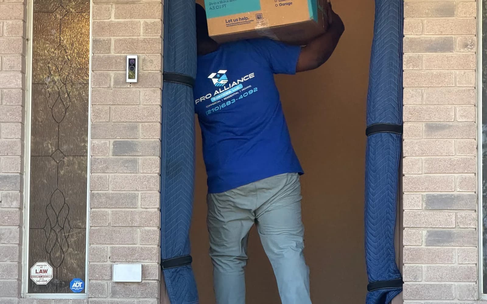 A Pro Alliance mover carrying a large labeled box through a front door with padded jamb covers
