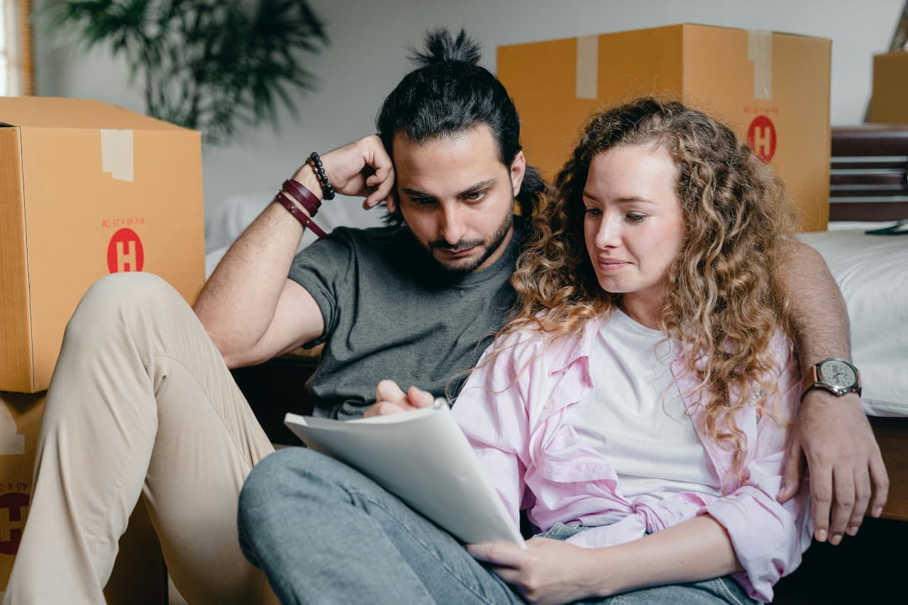 A young couple sitting on the floor surrounded by moving boxes, reviewing documents during their move.
