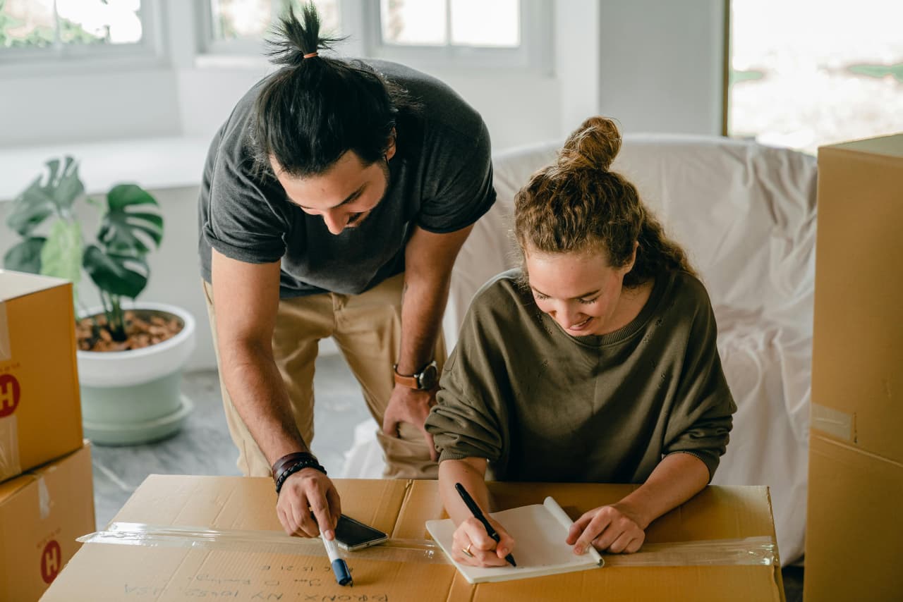 Man and woman labeling a cardboard moving box together while surrounded by other boxes in a bright living room.