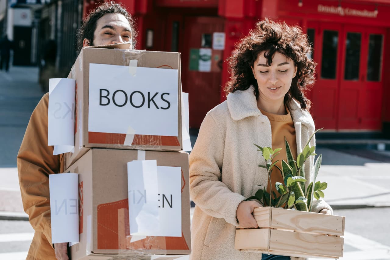 A young man carries stacked boxes labeled “BOOKS” while a young woman walks beside him holding a crate with a green houseplant, both smiling as they cross a city street during a move.
