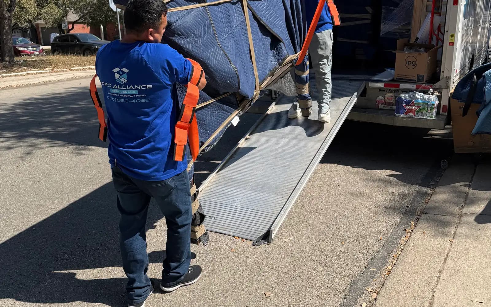 Two Pro Alliance crew members roll a strapped, blanket-wrapped item up a ramp into a box truck