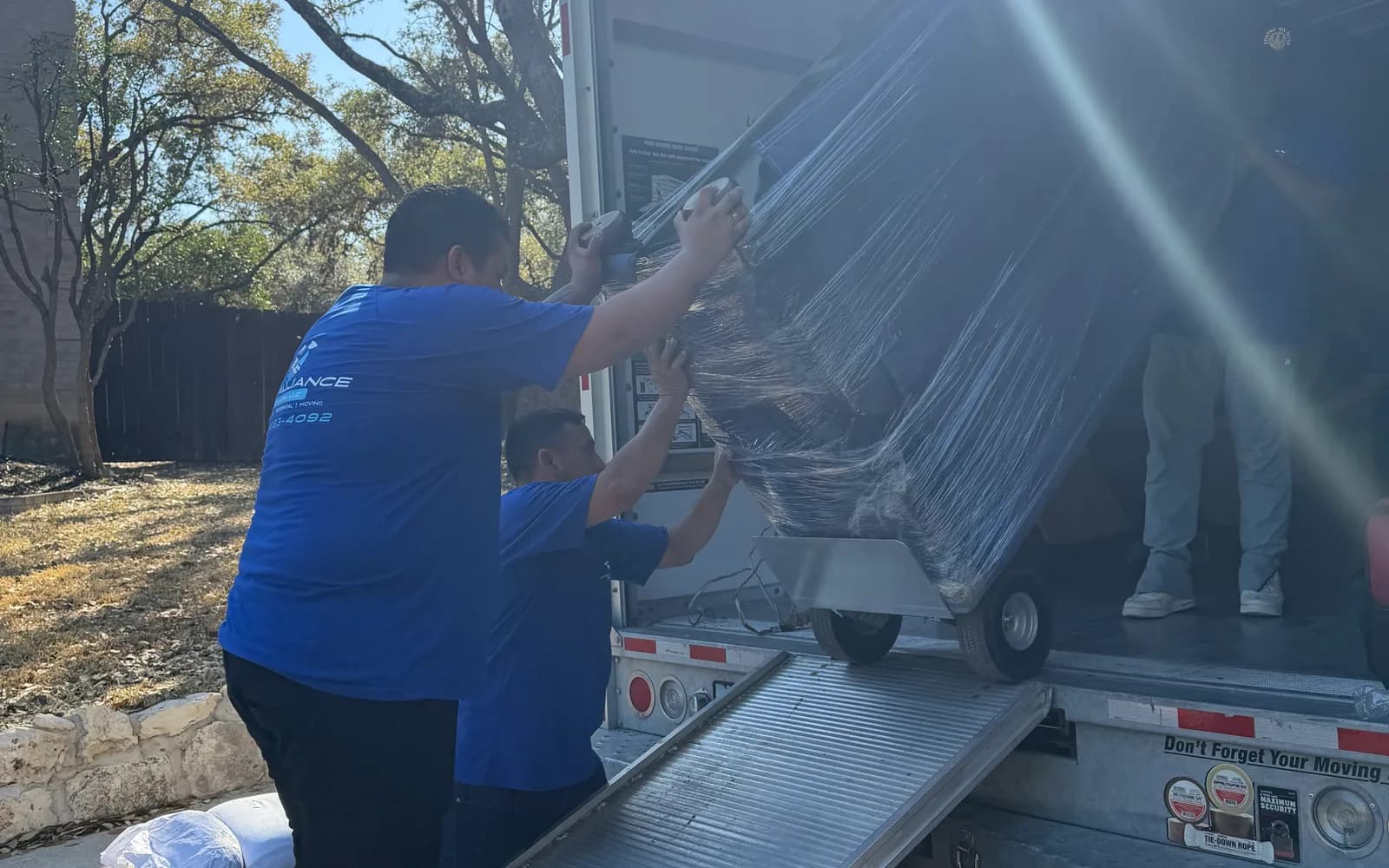 Pro Alliance crew members push a shrink-wrapped item on a dolly up a truck ramp in bright sunlight