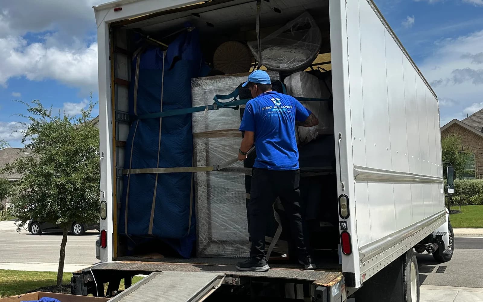 A Pro Alliance mover works inside a loaded box truck with the ramp down under a blue sky