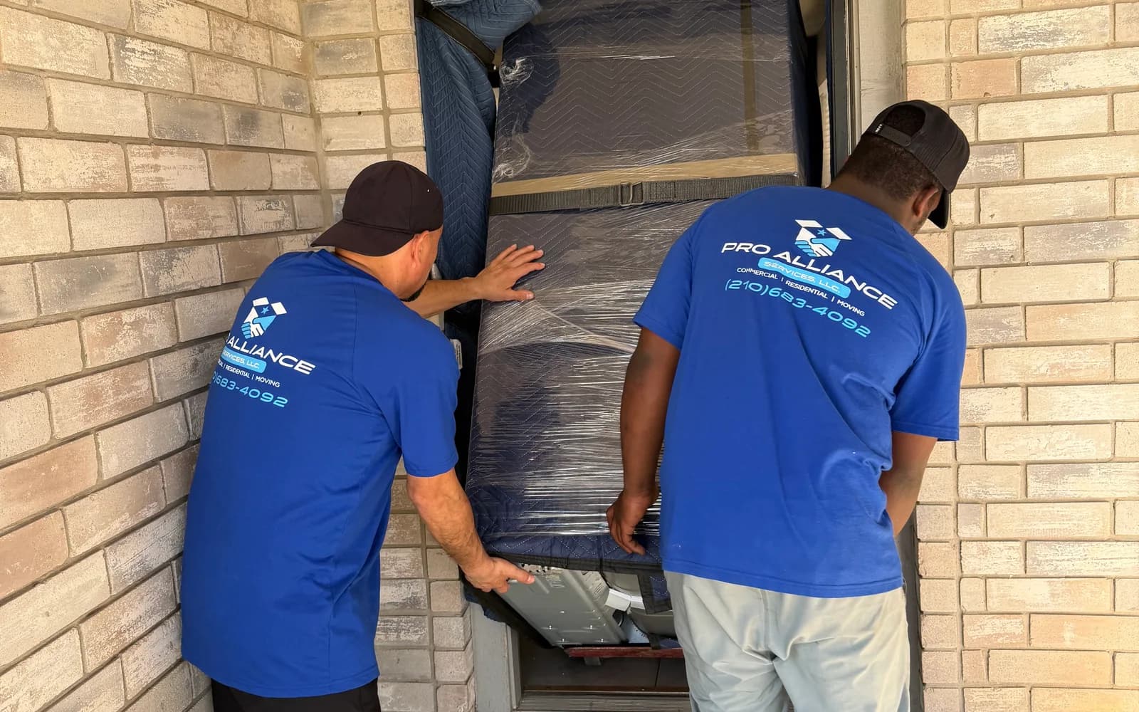 Two Pro Alliance crew members guide a wrapped mattress set through a padded entry door