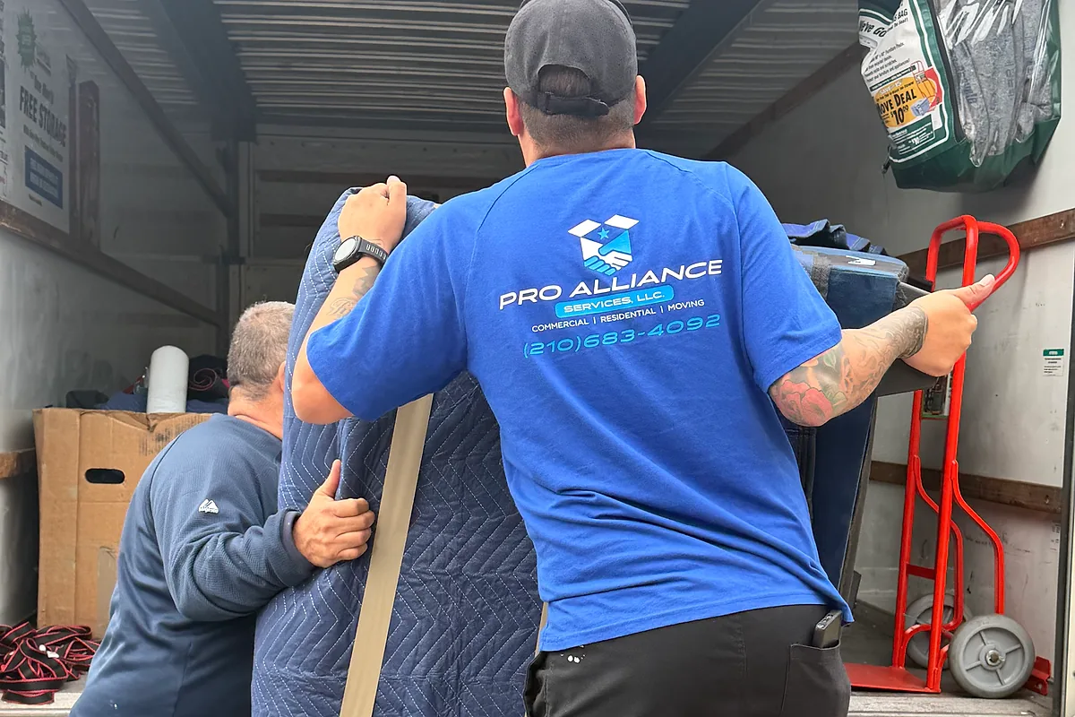 Two Pro Alliance movers carrying a blanket-wrapped piece up a loading ramp