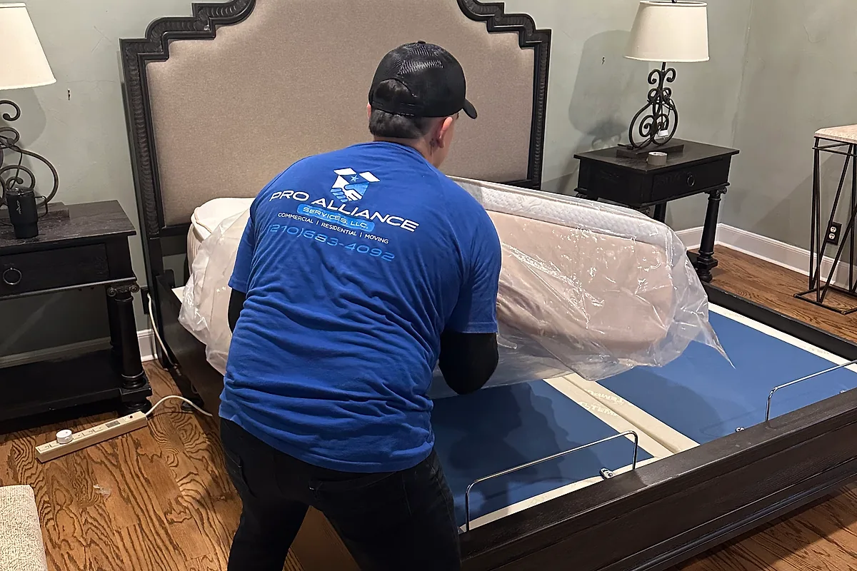 Pro Alliance mover wrapping a mattress in protective plastic beside a disassembled bed frame