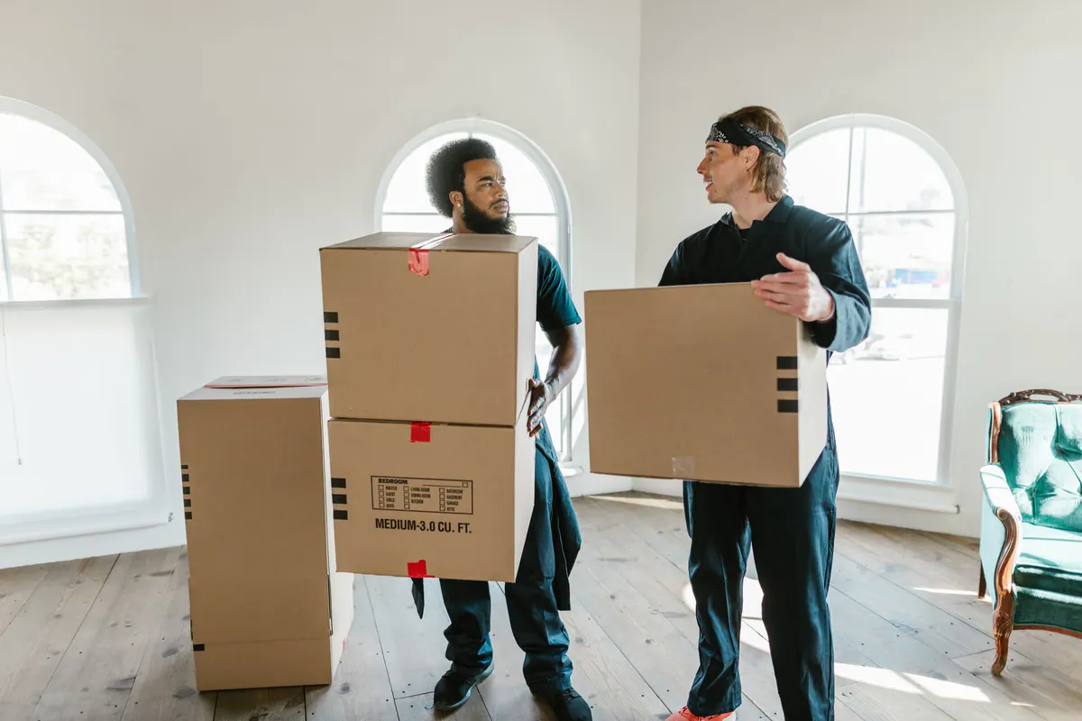 Two people carrying moving boxes through a bright room