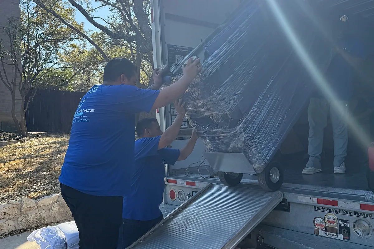 Pro Alliance movers bringing a wrapped item down a truck ramp on a tree-lined residential street