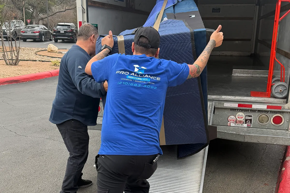 Two Pro Alliance movers guiding a blanket-wrapped piece up a truck ramp
