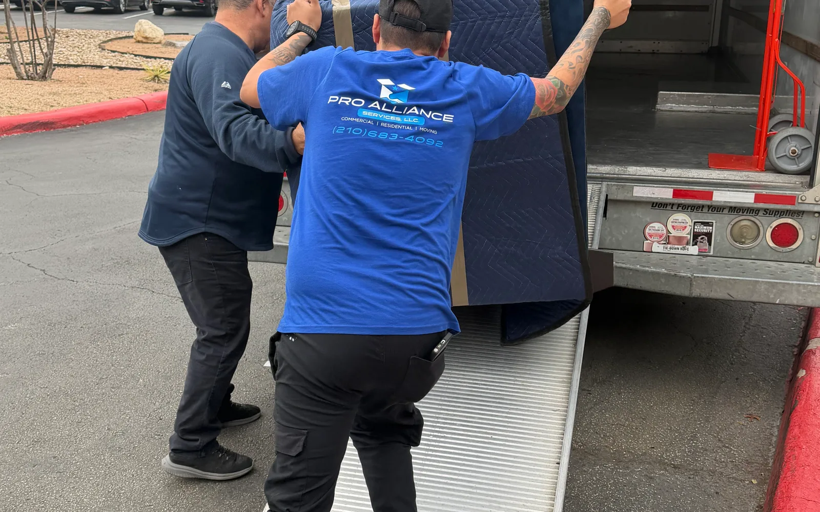Two Pro Alliance crew members seen from behind push a blanket-wrapped item up a walk ramp into a truck