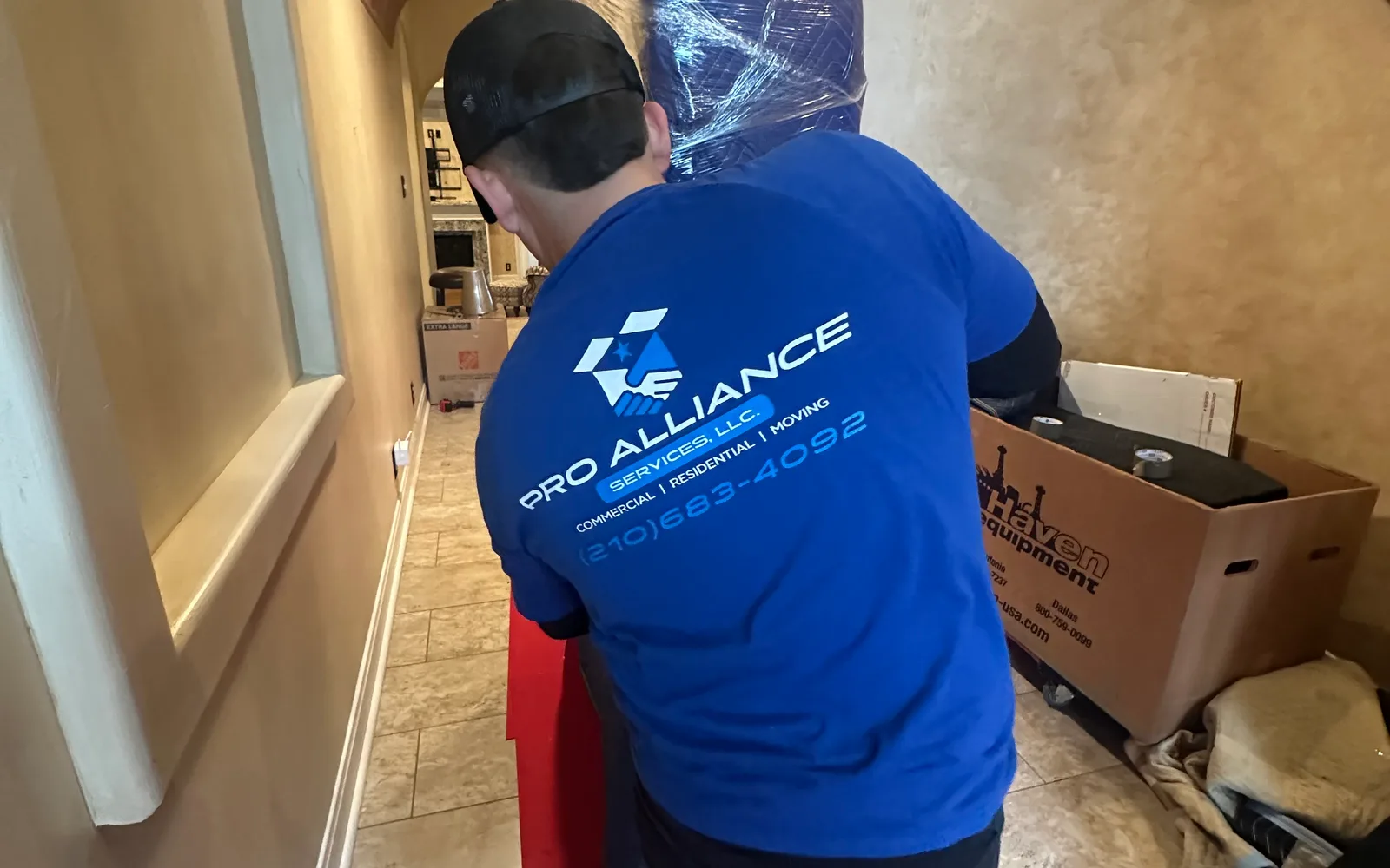 A Pro Alliance mover guides a plastic-wrapped item along a tiled hallway with a floor runner in place