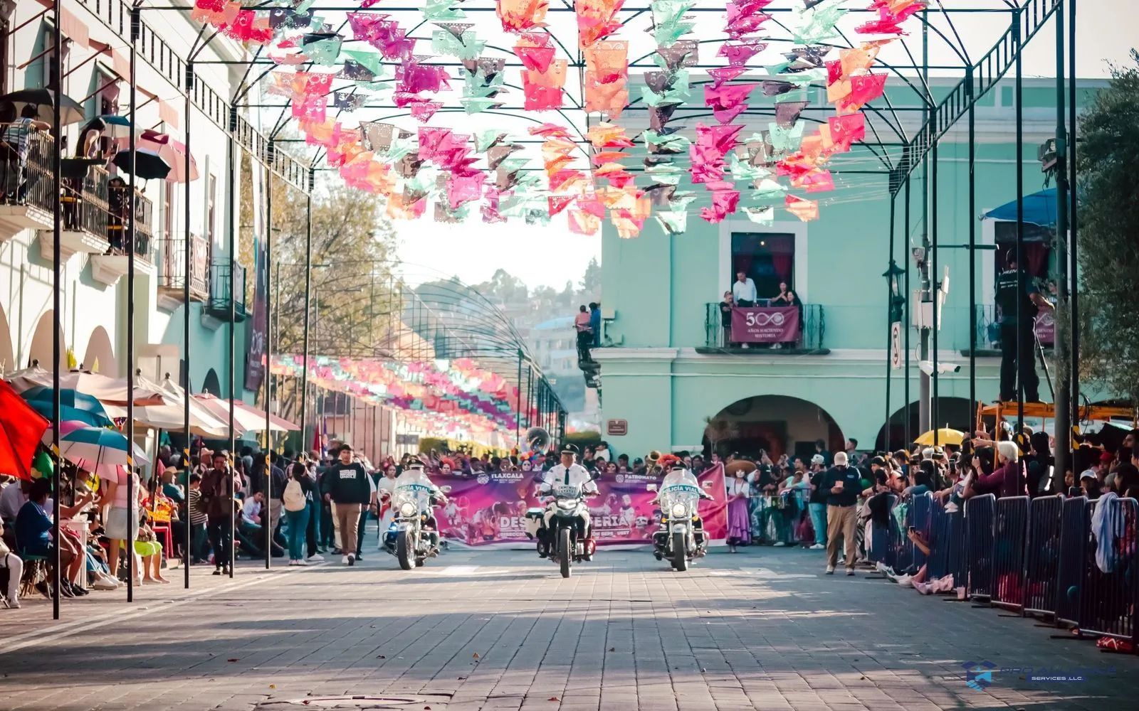 A street parade under strings of colorful paper banners, with crowds behind barricades