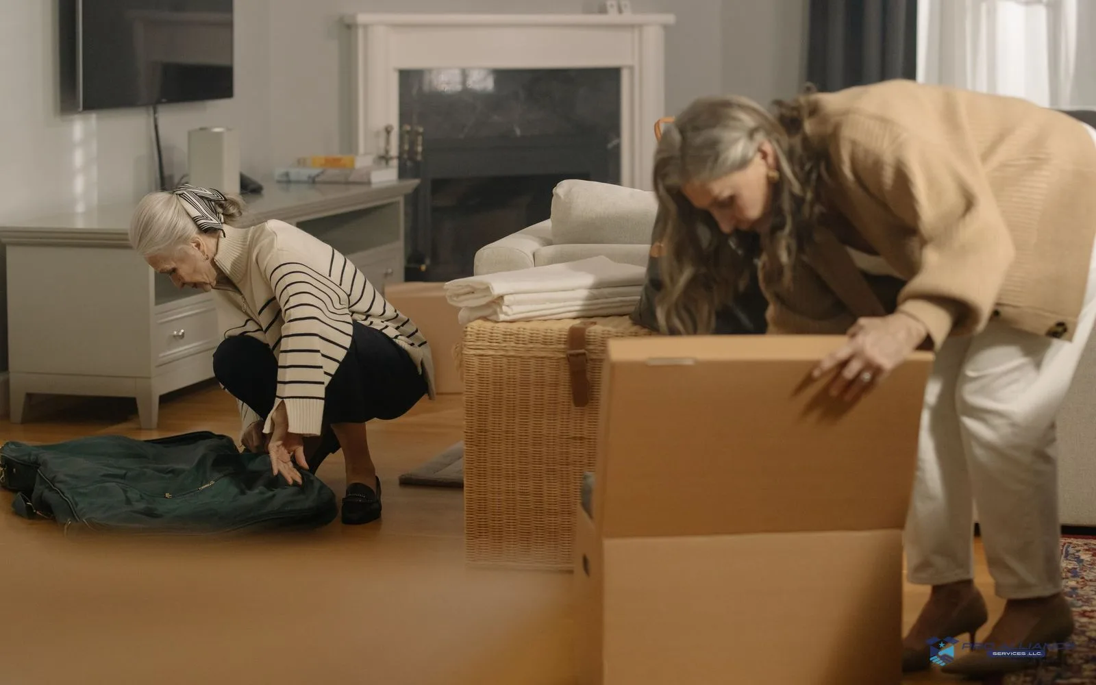An older woman zipping a garment bag while a younger woman packs a cardboard box in a living room