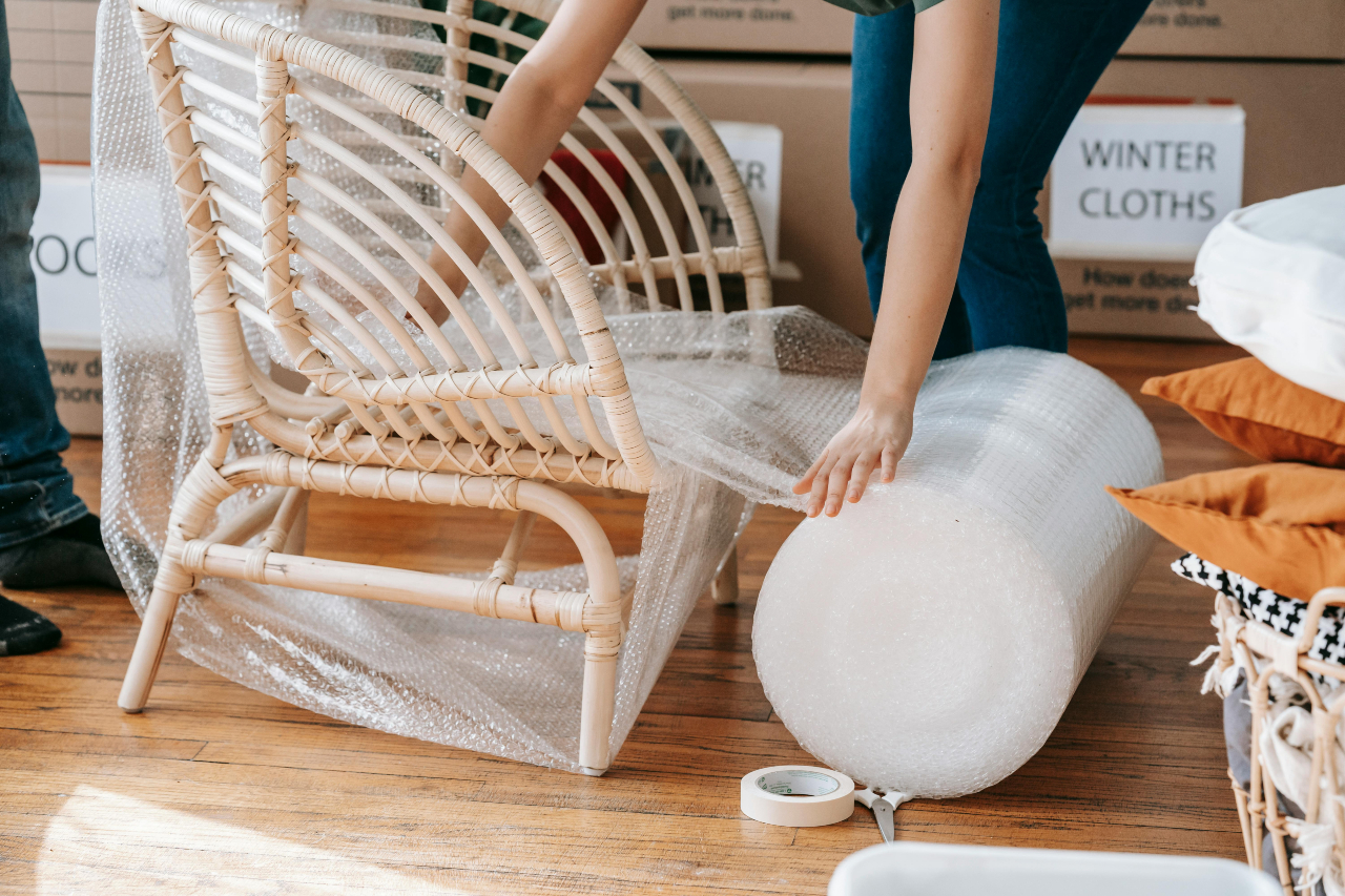Person carefully wrapping a wicker chair with bubble wrap, preparing furniture for safe transport during a move.