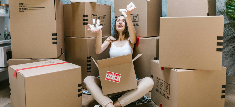 Happy young woman playing with packing peanuts among boxes while preparing her new space after leaving her parents’ home for the first time.