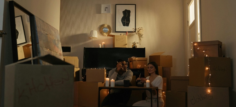 Couple sitting on the floor eating dinner with candles among moving boxes in their new home.