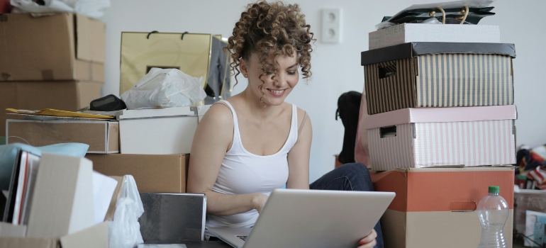 A woman is sitting on the floor with her laptop around boxes.