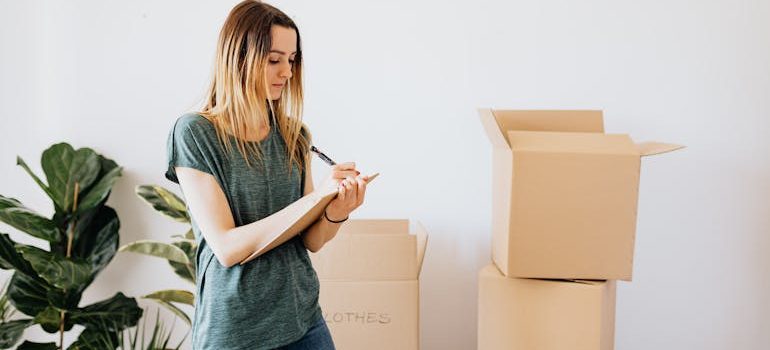 A young woman stands amid unpacked boxes, holding a pen and a notepad while following her checklist to help her with the same-day move in UValde.